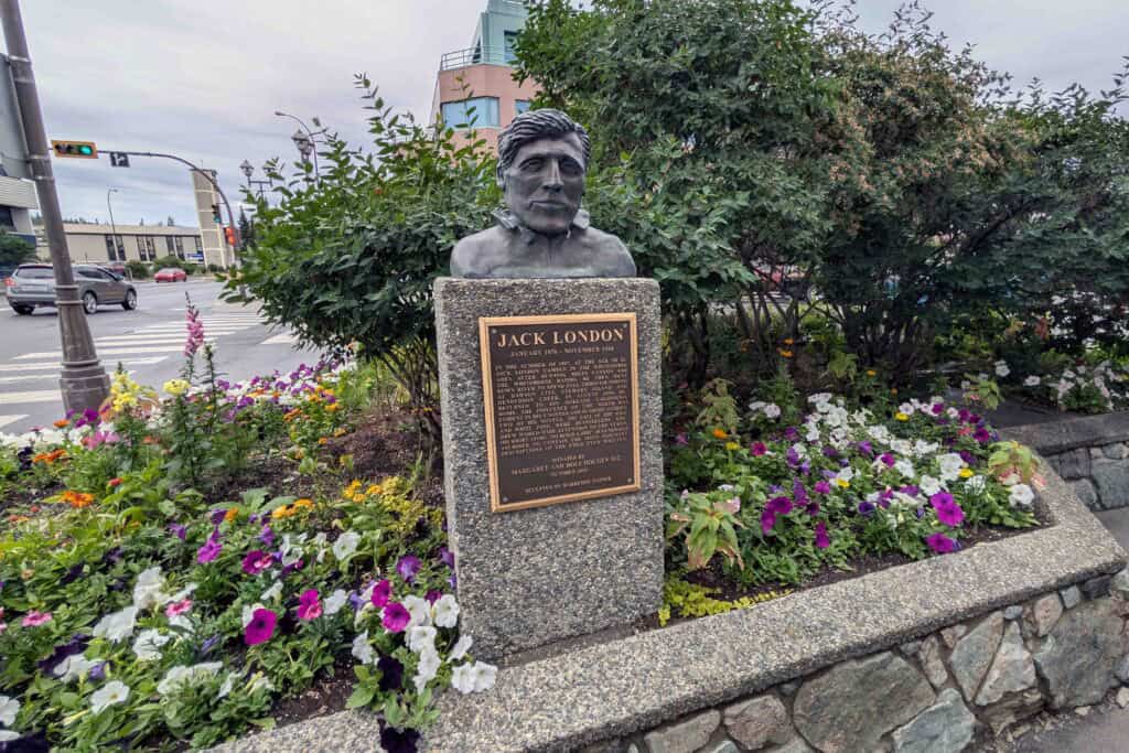 Jack London memorial in downtown Whitehorse, featuring a bronze bust on a stone pedestal with a plaque, surrounded by colourful flower beds at a street corner.