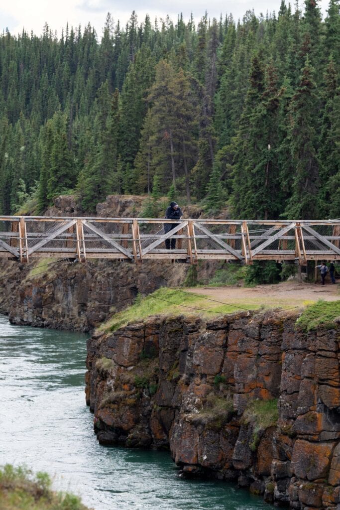 Wooden suspension bridge spanning Miles Canyon near Whitehorse, with steep rocky canyon walls, turquoise river water below, and evergreen forest in the background.