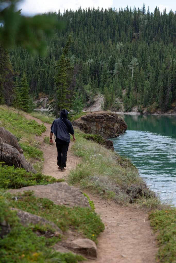 Person walking along a narrow dirt trail beside the Yukon River at Miles Canyon, with rocky cliffs, calm turquoise water, and dense evergreen forest lining the canyon.