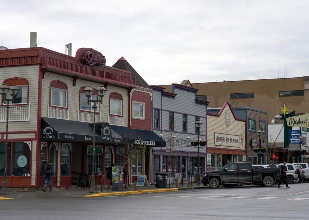 Colourful historic storefronts lining a main street in downtown Whitehorse, with locally owned shops, restaurants, parked vehicles, and pedestrians on an overcast day.