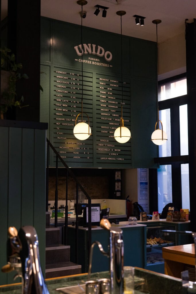 Interior of Unido Panama Coffee Roasters with a large green menu board, hanging globe lights, and a counter displaying pastries and coffee equipment.