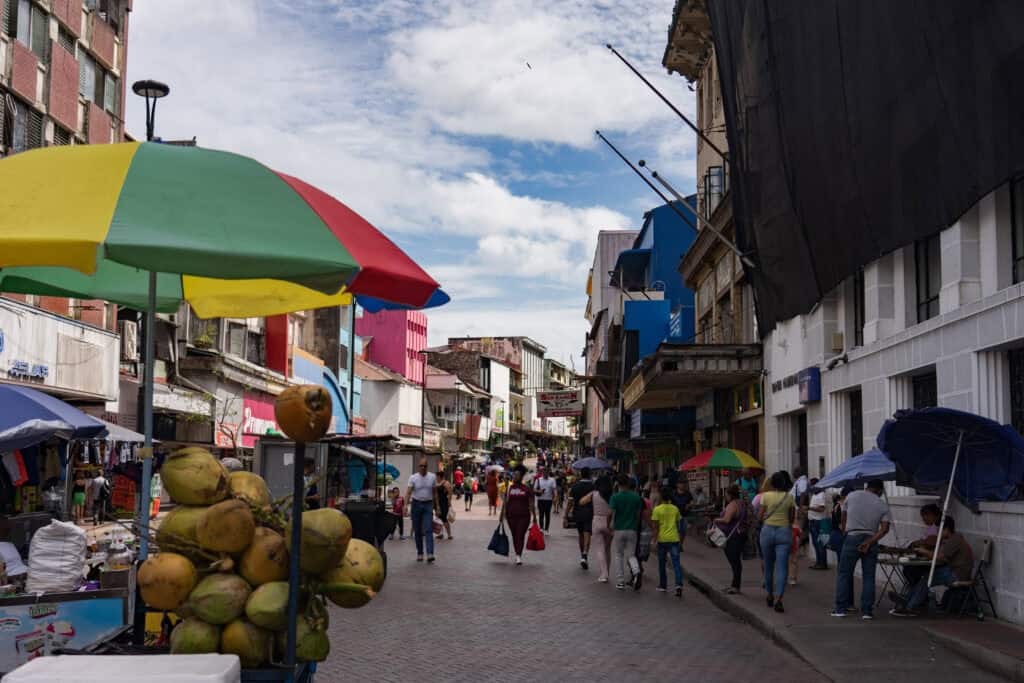 A busy pedestrian shopping street in Panama City lined with colorful storefronts and market stalls, where people walk beneath umbrellas and a vendor displays fresh coconuts under a bright green, yellow, and red canopy.