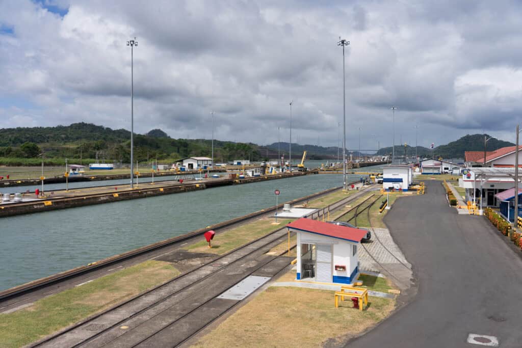 A wide view of the Miraflores Panama Canal locks with parallel waterways, rail tracks, and service buildings stretching into the distance under a cloudy sky.
