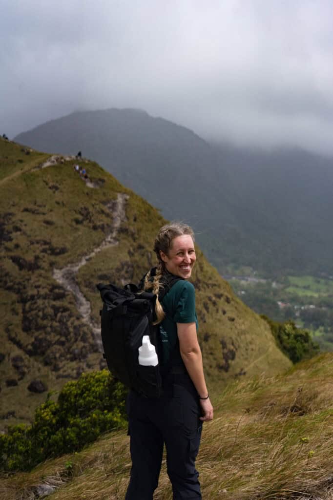 A smiling woman with braided hair and a large black backpack stands on a grassy mountain ridge, looking back at the camera, with misty hills and a winding trail behind her.