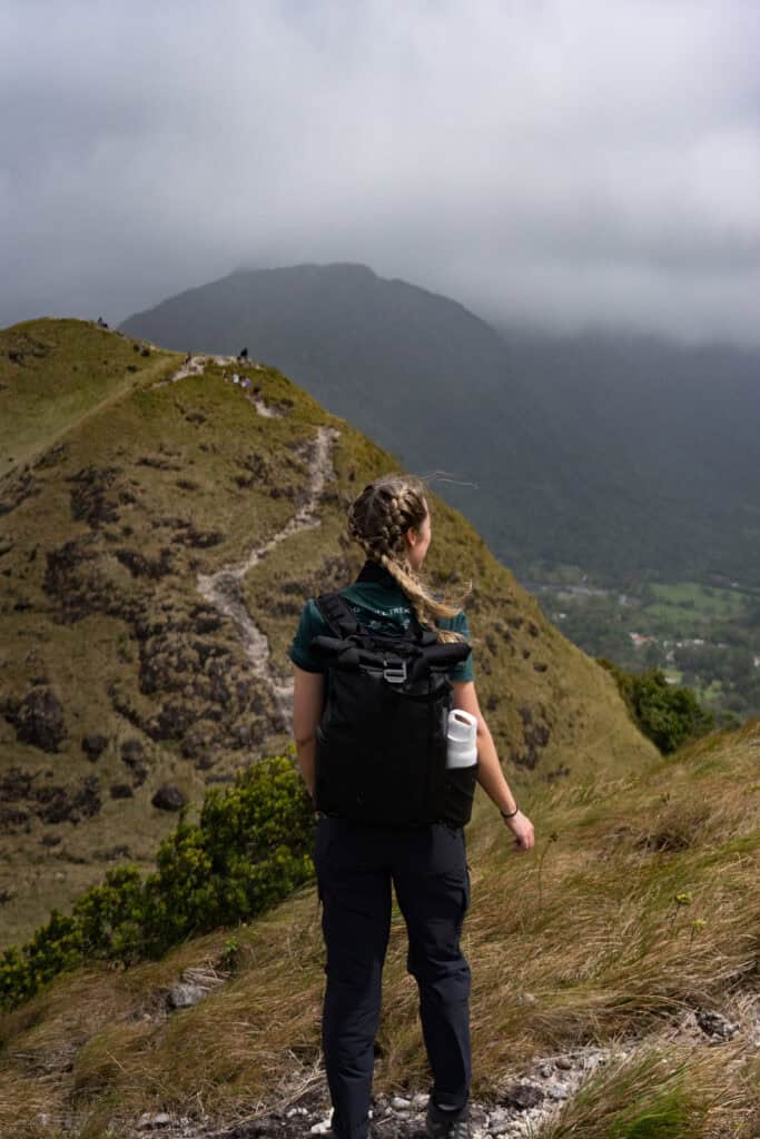 A woman with braided hair and a black backpack stands on a narrow, grassy mountain ridge, looking out toward mist-covered hills as a winding trail and small hikers stretch along the ridgeline ahead.