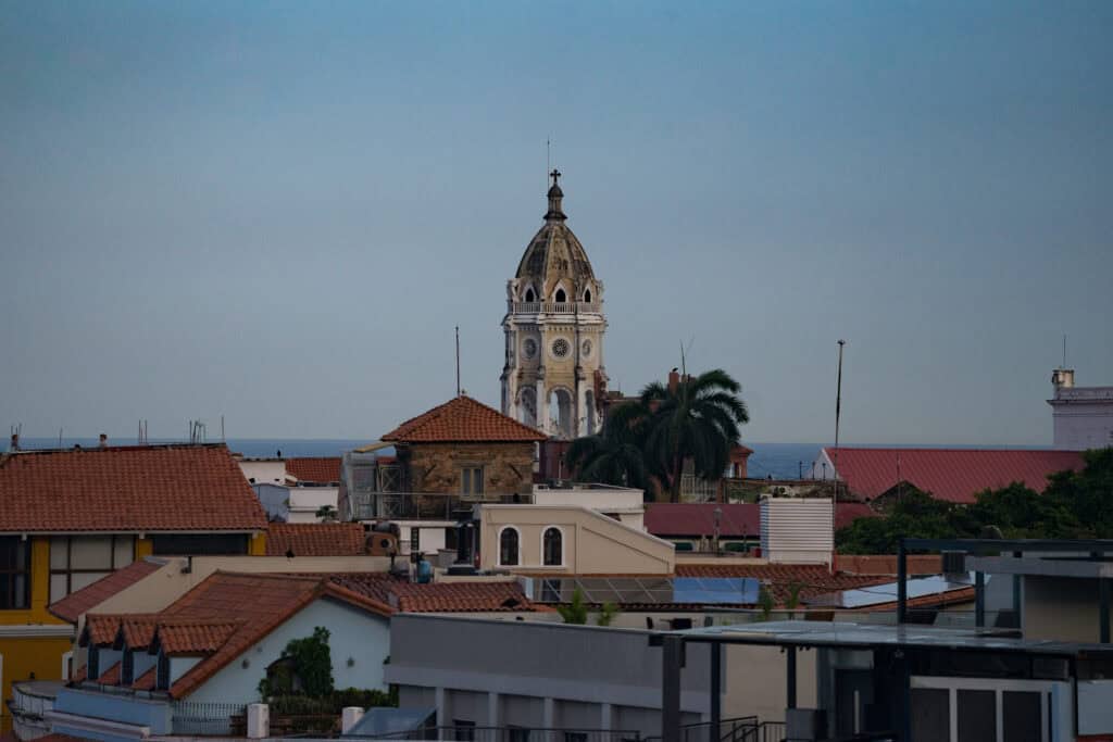 View over red-tiled rooftops in Panama City’s historic district, with a white cathedral bell tower rising above the buildings and the ocean visible in the background.