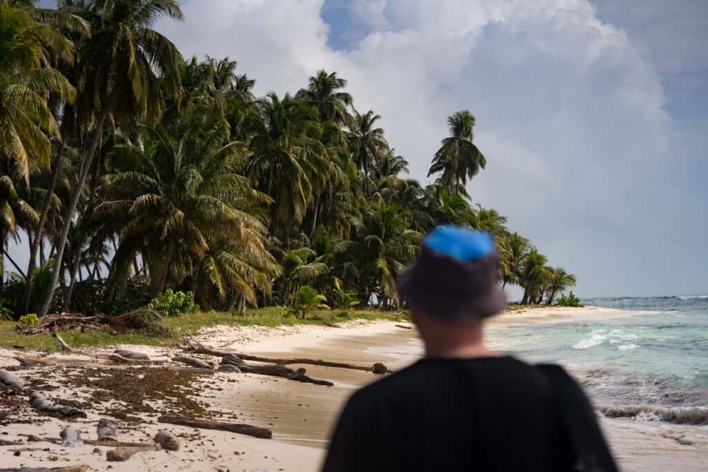 A man wearing a hat walking along a beach on El Porvenir, Guna Yala, with palm trees ahead of him.