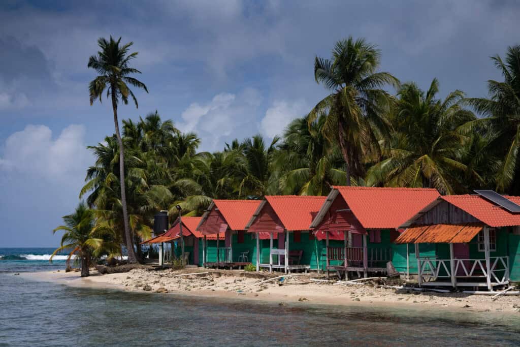 A row of small, brightly painted cabins with red roofs sits along a sandy shoreline lined with tall palm trees, with the ocean visible just beyond the beach.