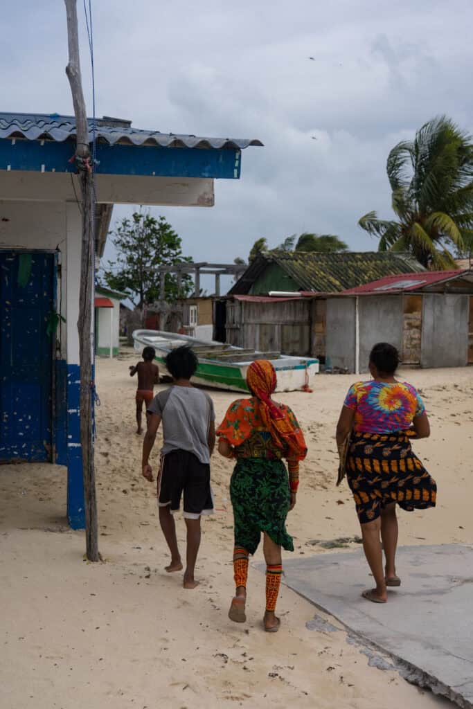 Three people walking along a sandy street in a Guna Yala island community, with simple homes, boats, and palm trees in the background.