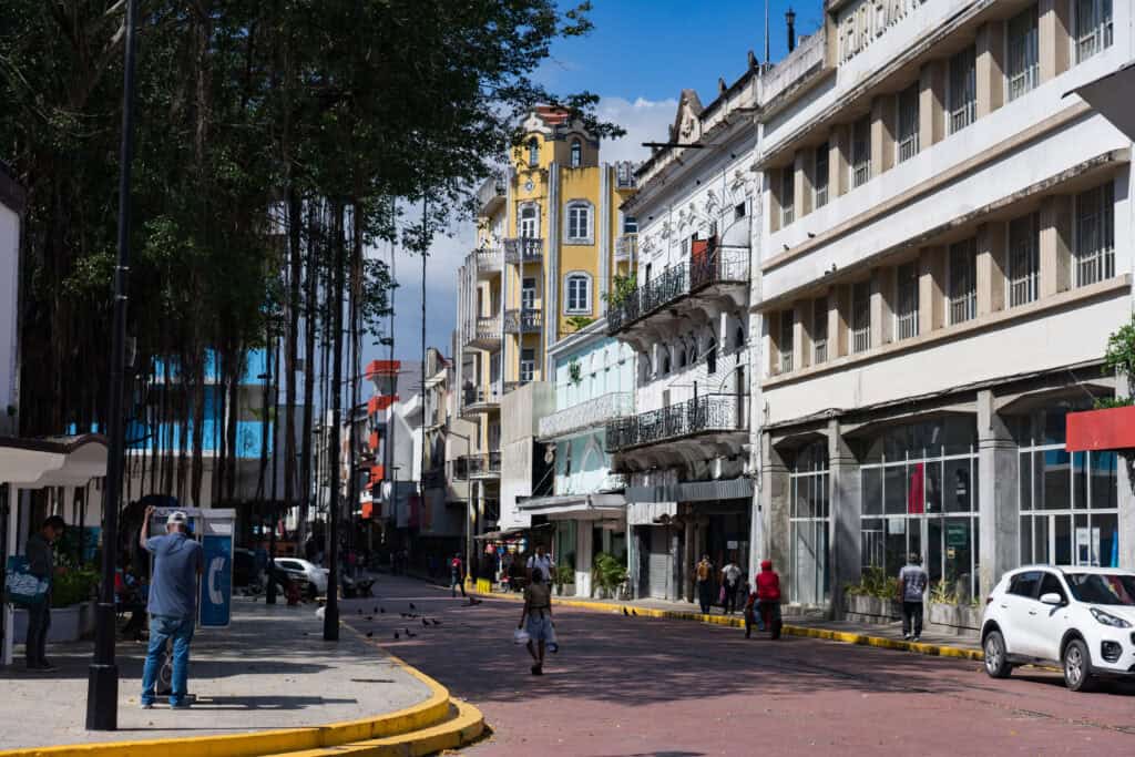 A sunny street scene in Panama City with colorful historic buildings and wrought-iron balconies lining the road, pedestrians walking along the brick-paved street, and tall trees casting shade over the sidewalk.