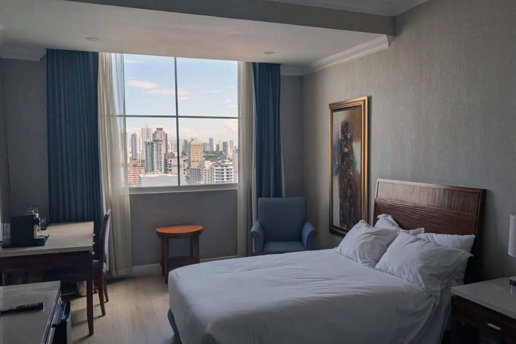 Bright hotel room in Panama City with a large bed, desk, armchair, and floor to ceiling window overlooking a skyline of high rise buildings.