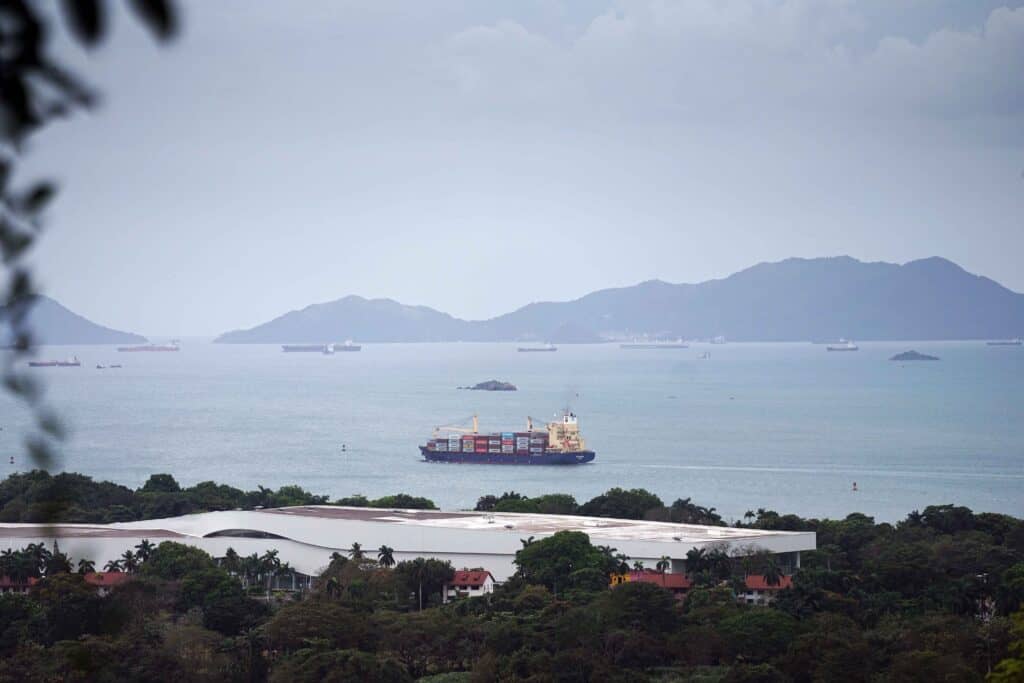Container ship sailing in the Bay of Panama with several cargo vessels in the distance and green hills rising beyond the water.