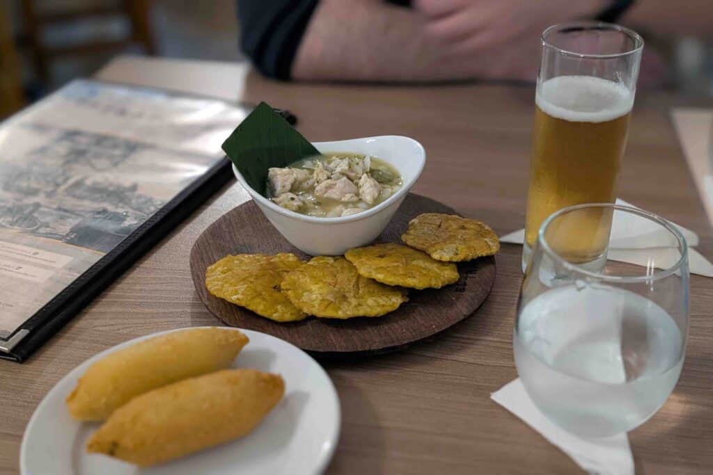 Traditional Panamanian meal on a wooden table, including a bowl of ceviche, patacones on a wooden plate, fried Carimañolas, and a glass of beer.