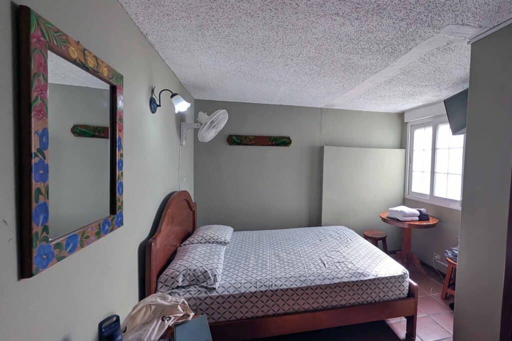 Modest guest room in El Valle de Antón with a double bed, small round table by the window, tiled floor, and a colorful framed mirror on the wall.
