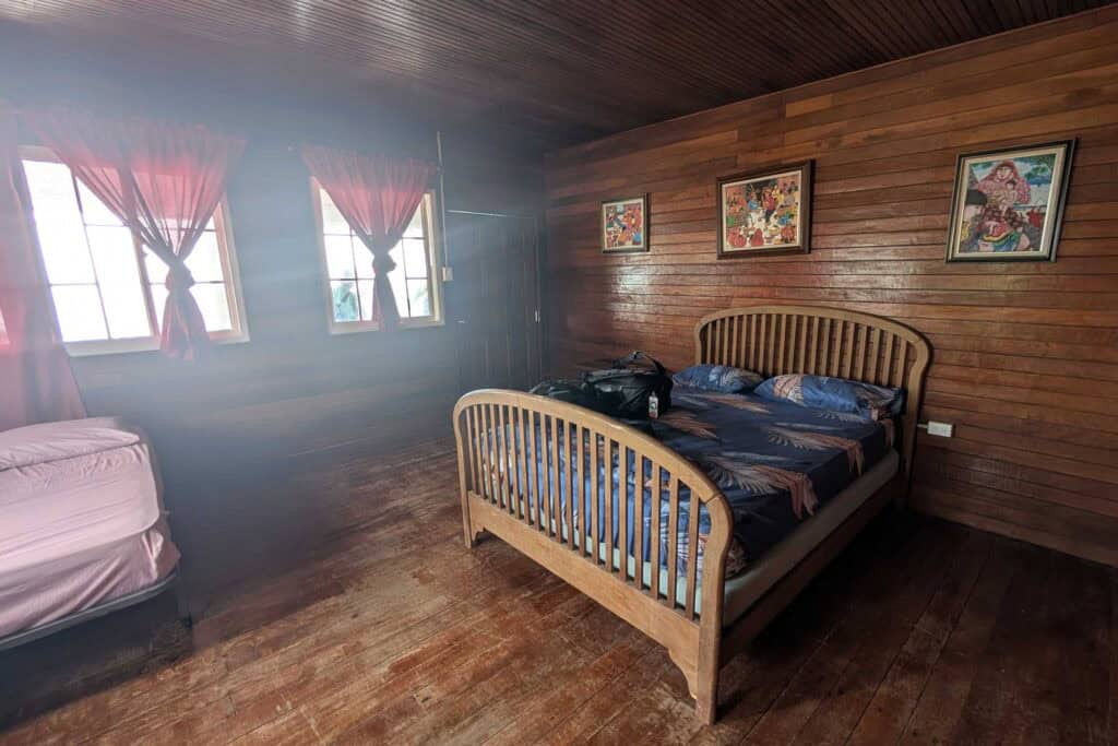 Simple wooden guest room in Guna Yala with wood-paneled walls, a double bed, red curtains over two windows, and framed artwork above the headboard.