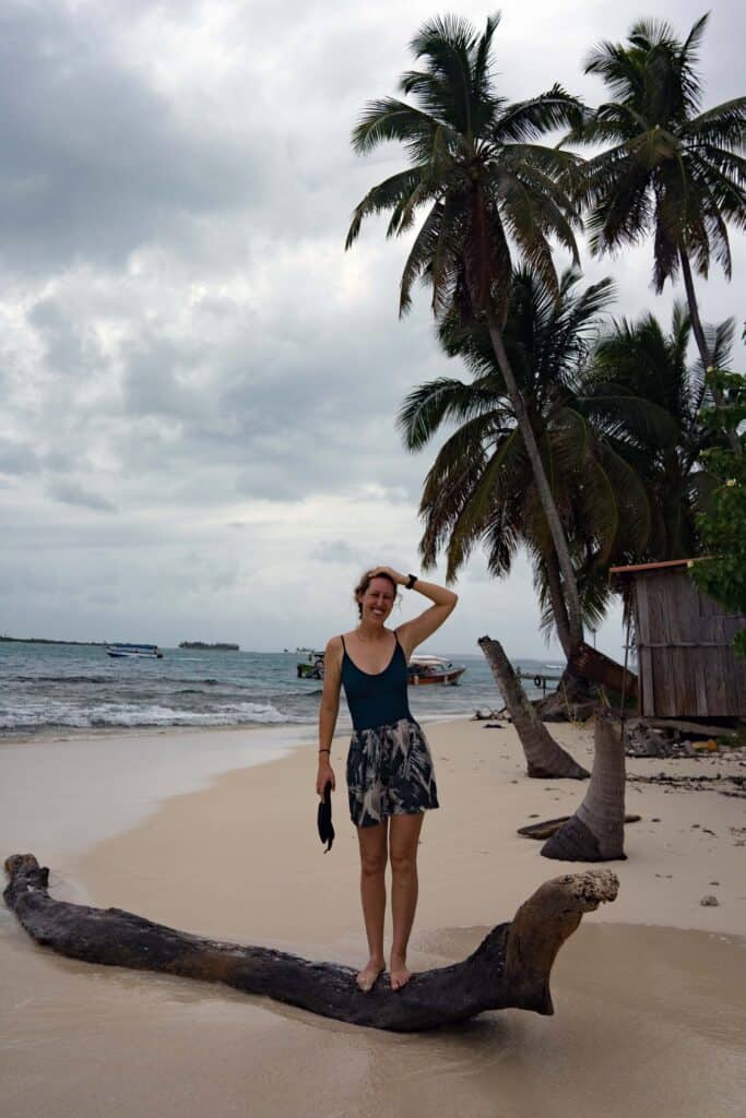 Woman standing on a driftwood log on a sandy beach in Guna Yala, Panama, with palm trees, small boats, and Caribbean coastline in the background.
