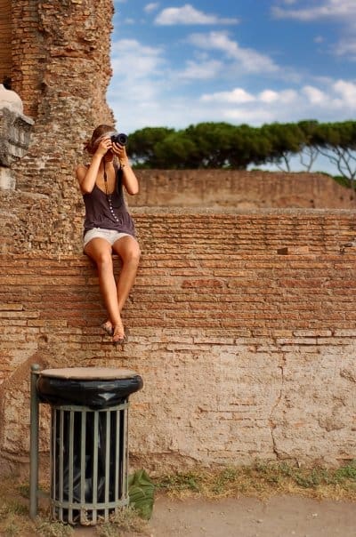 Travel blogger sitting on a brick wall taking photos at ancient Roman ruins in Rome, Italy.