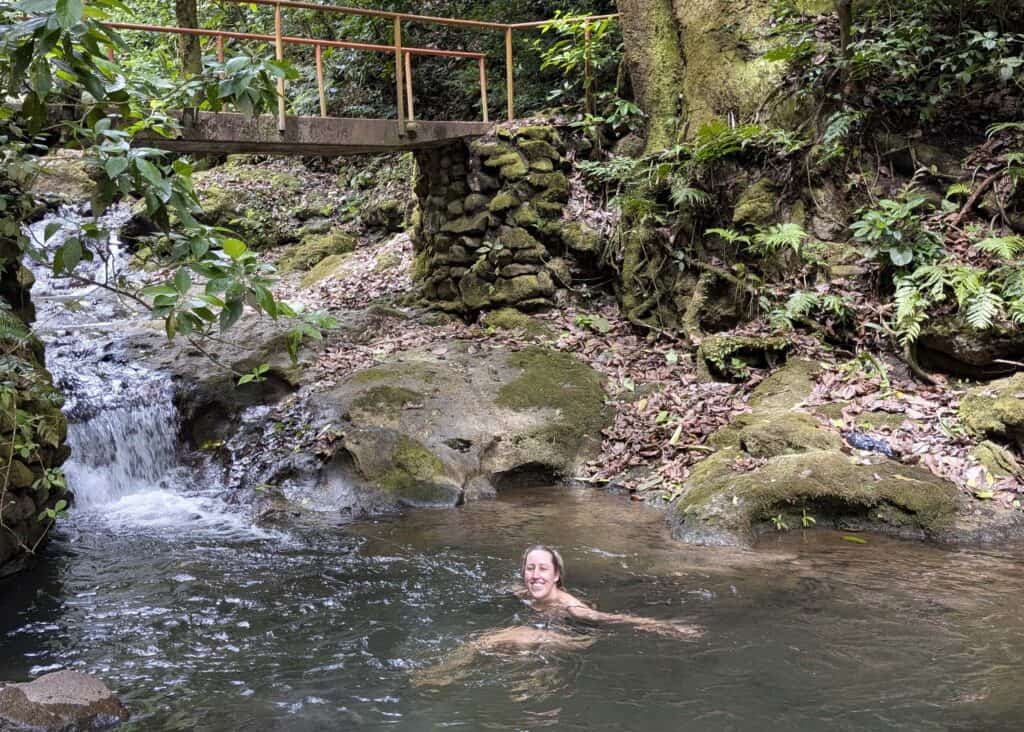 Person swimming in a natural pool beneath a small waterfall on La India Dormida trail, with a wooden bridge and lush jungle surroundings in El Valle de Antón