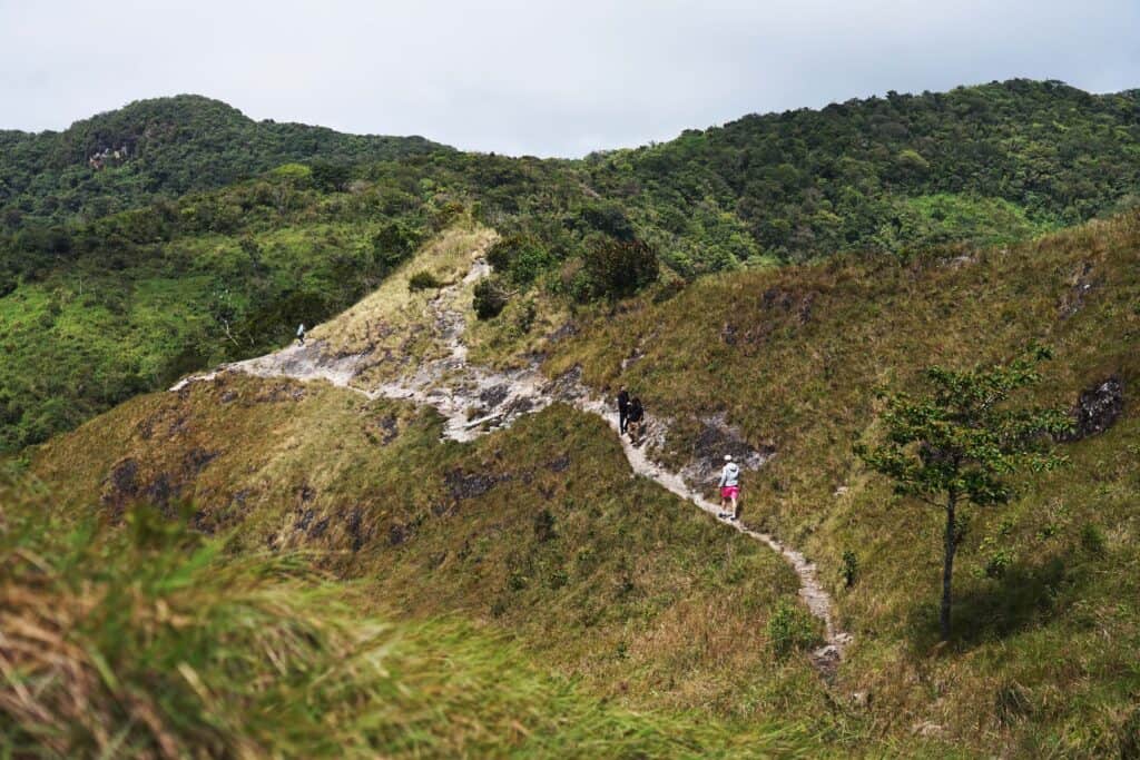 Hikers walking along a narrow ridge trail on La India Dormida with grassy slopes and rolling hills in El Valle de Antón.