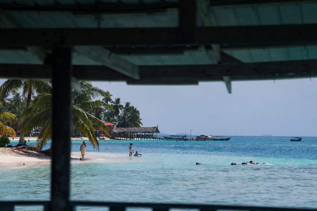 View of a beach in Guna Yala, Panama, with clear shallow Caribbean water, people swimming and snorkeling, palm trees along the shore, and wooden docks with small boats in the distance.