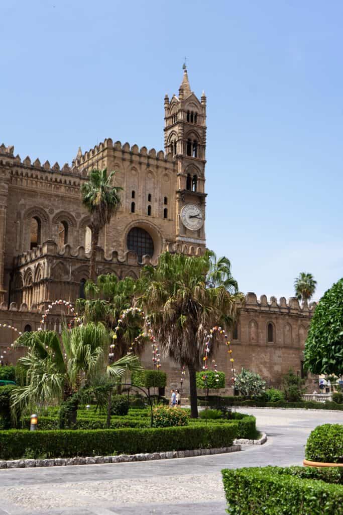 View of Palermo Cathedral (Cattedrale di Palermo) in Palermo, Sicily, with its ornate Norman-style bell tower and clock rising above palm trees and landscaped gardens in the square below.