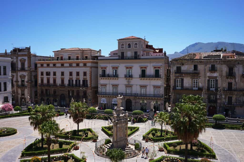 View over a landscaped square in Palermo with palm trees, statues, and geometric gardens, surrounded by historic buildings and backed by mountains under a clear blue sky.