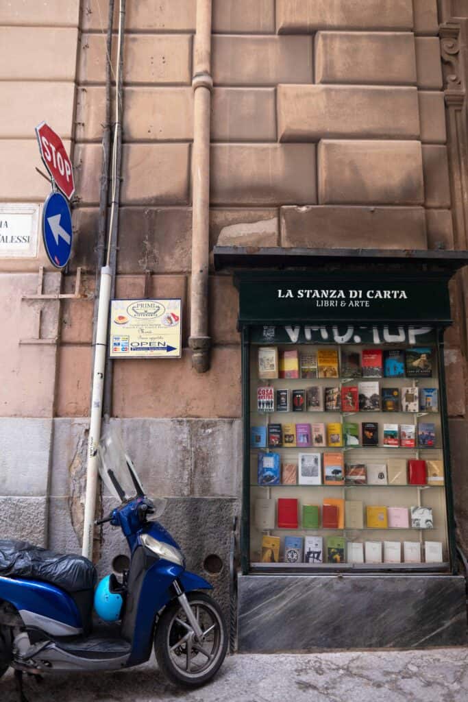 Street scene in Palermo’s historic center showing a blue scooter parked beside a bookstore window display labeled “La Stanza di Carta – Libri & Arte,” with books visible behind the glass and weathered stone walls and street signs above.