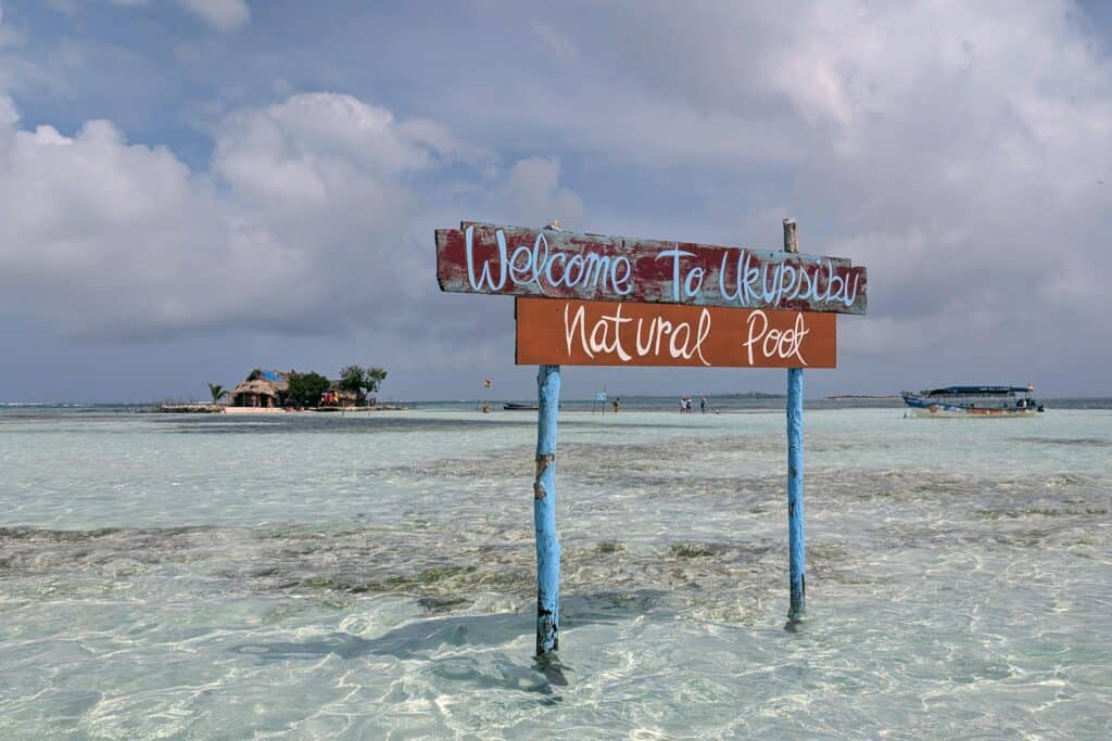 Sign reading “Welcome to Ukupseni Natural Pool” standing in shallow clear water in Guna Yala, Panama, with a sandbank, small boats, and a tiny island in the background.