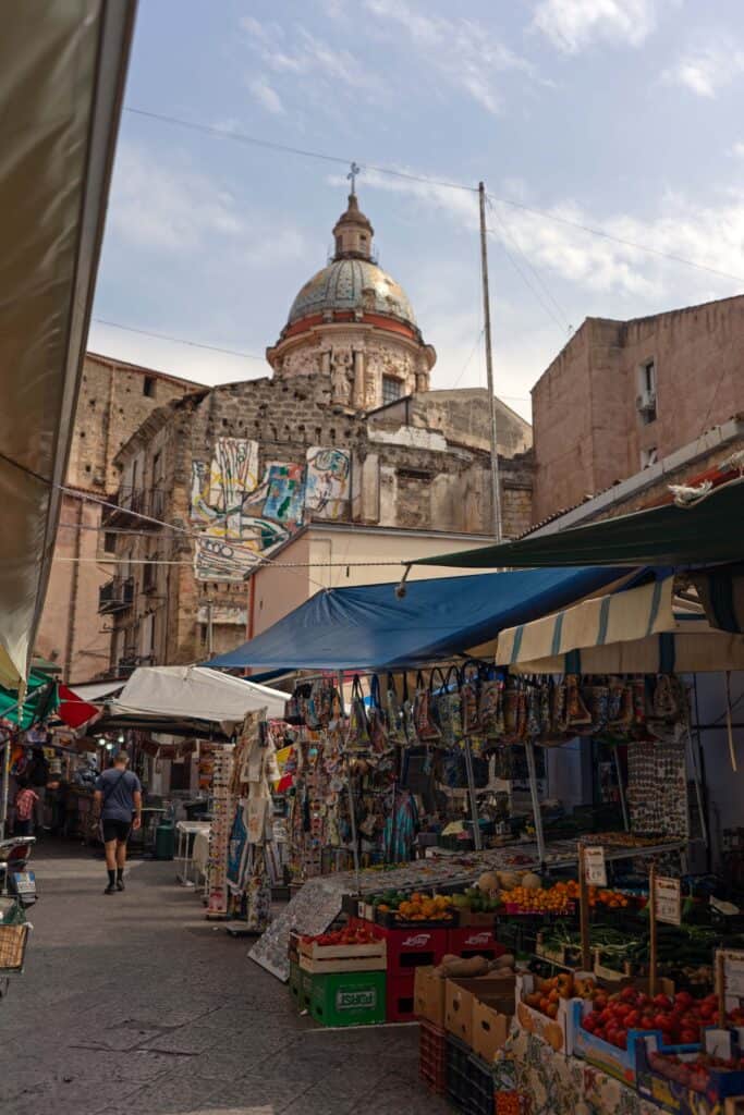 Street market stall selling fresh produce in Ballarò Market in Palermo, Sicily, with colorful fruits and vegetables under striped awnings and the dome of a historic church rising above the surrounding buildings.