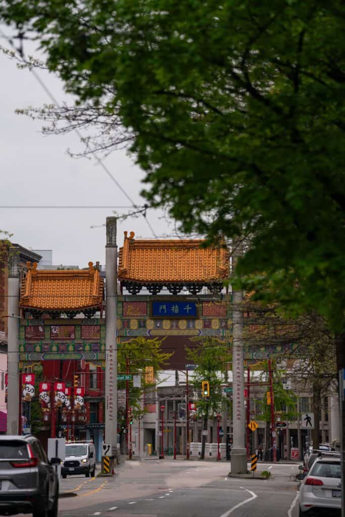 A wider street view of Vancouver’s Chinatown shows the Millennium Gate in the distance beneath overhanging green tree branches.