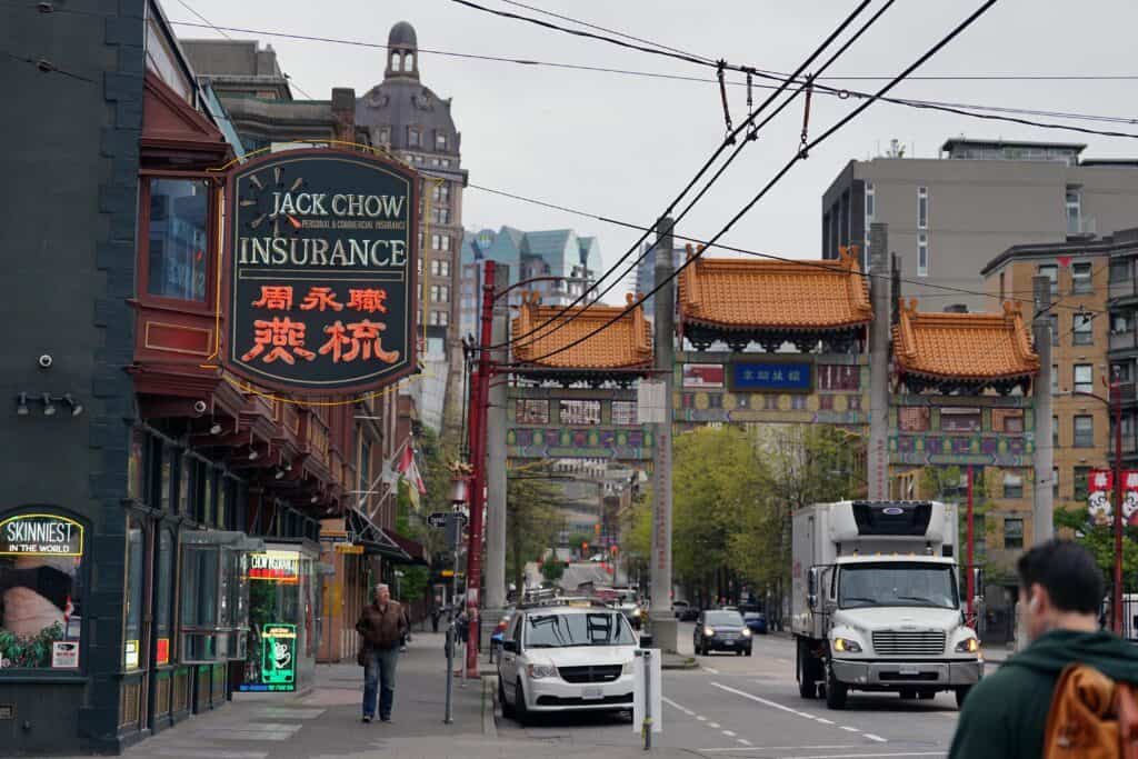 A street view of Vancouver’s Chinatown shows the Millennium Gate in the distance alongside the historic neon “Jack Chow Insurance” sign and passing traffic.