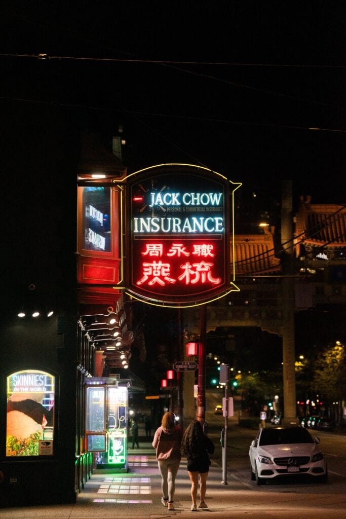 Neon signs glow along a quiet street in Vancouver’s Chinatown at night as two people walk beneath the illuminated “Jack Chow Insurance” sign.
