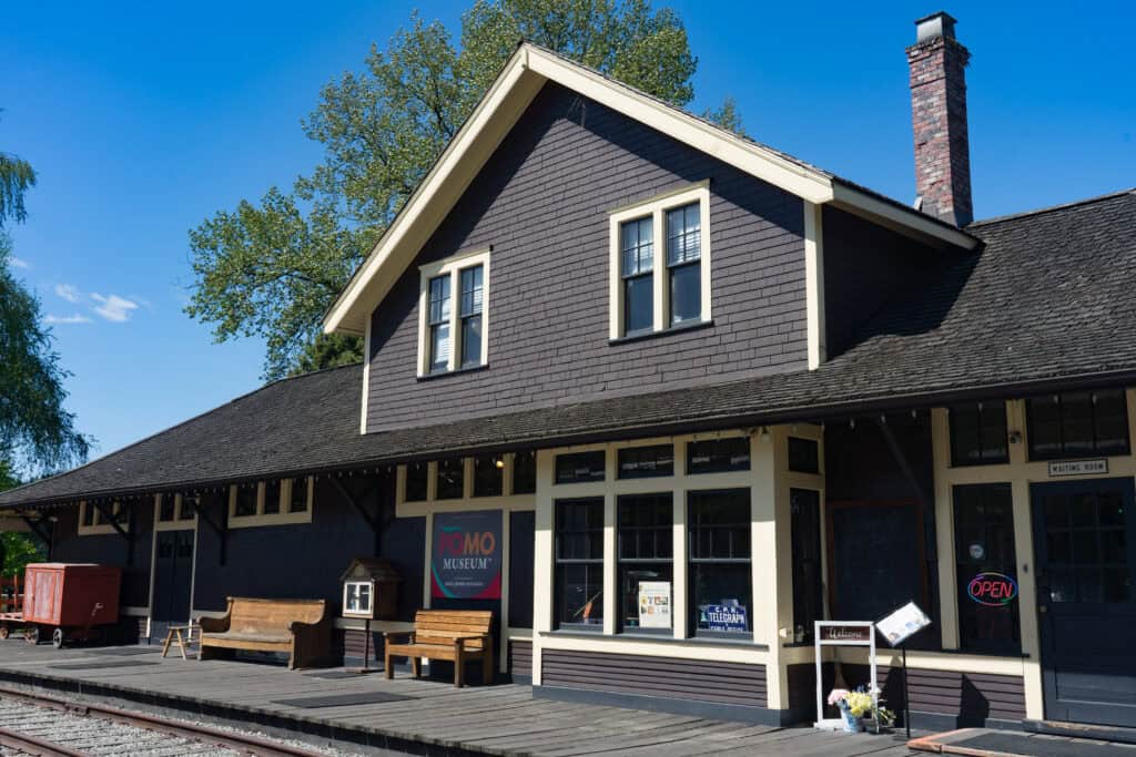 Exterior of the PoMo Museum housed in a restored historic CPR train station building, with dark siding, cream trim, wooden benches, and a sign welcoming visitors near the entrance.