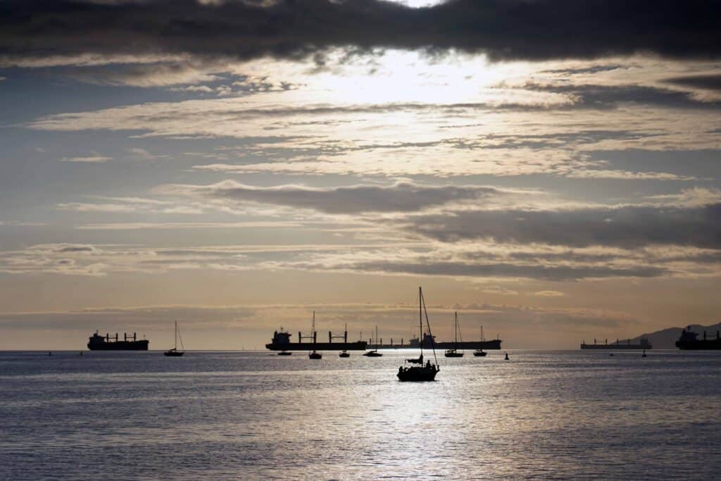Sailboats and cargo ships silhouetted on calm water at sunset in Vancouver, with soft golden light reflecting across the ocean and layered clouds stretching across the sky.