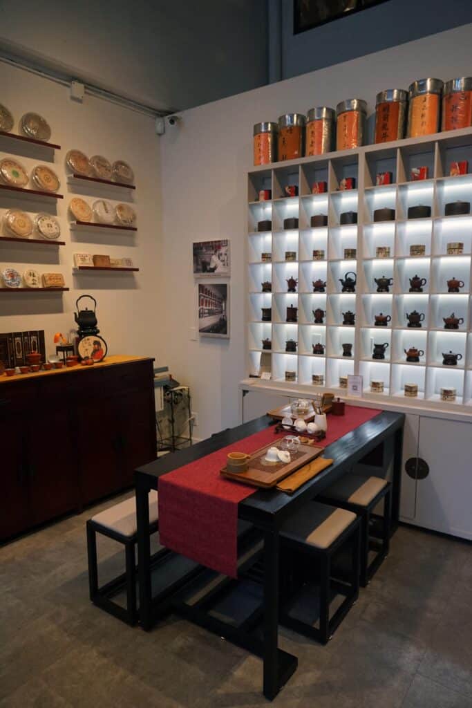 A tea tasting table sits inside a Chinatown tea shop lined with teapots, tea tins, and illuminated display shelves.