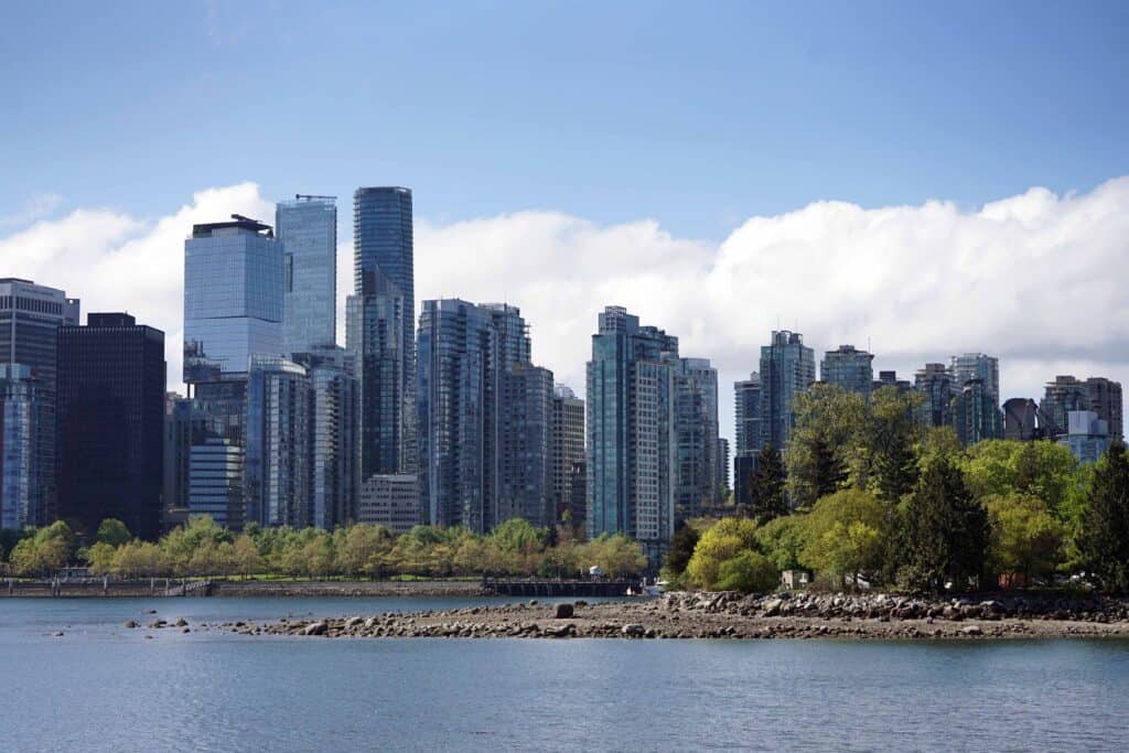 Vancouver skyline viewed from the waterfront, with modern high-rise buildings backed by low clouds and greenery along the shoreline.