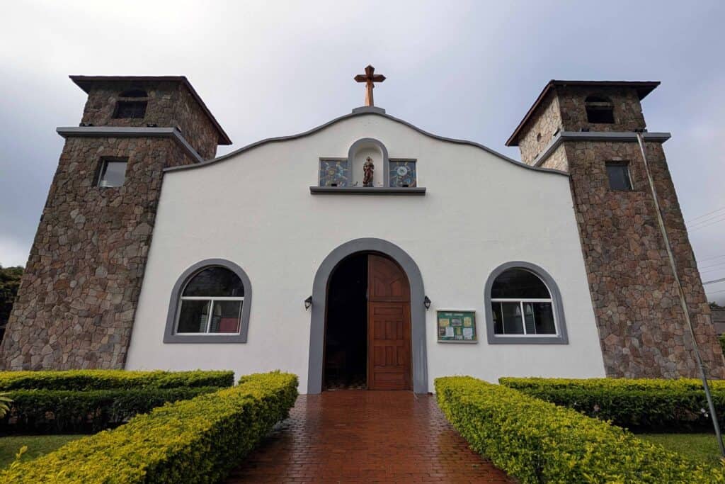Front view of Iglesia San José in El Valle de Anton, Panama, featuring stone bell towers, a white facade, and a brick walkway lined with trimmed hedges.