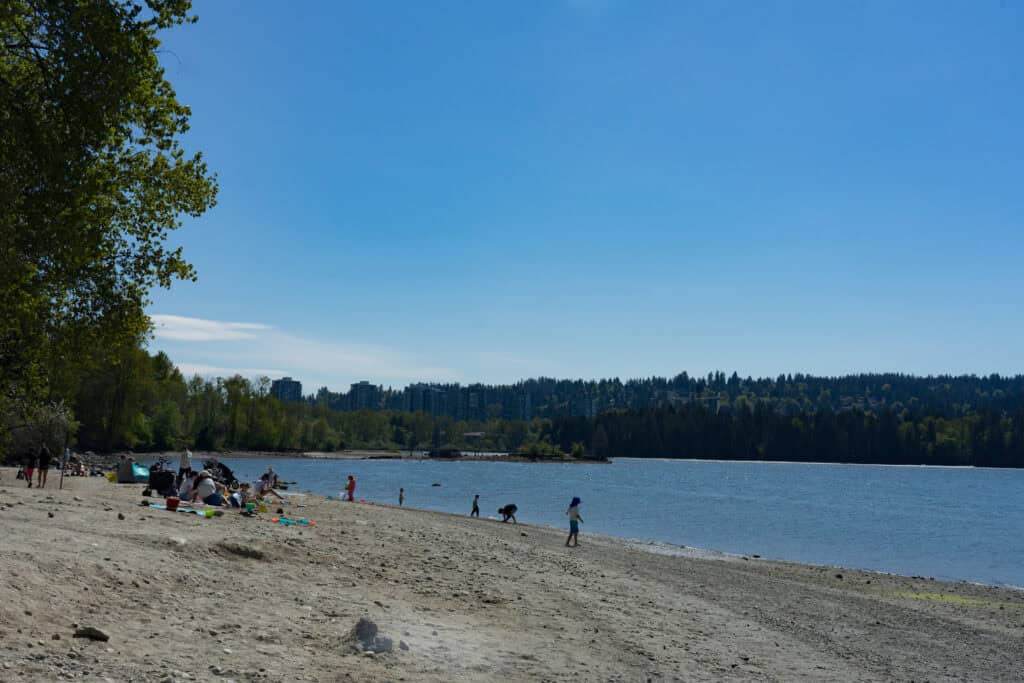 Sandy beach at Old Orchard Park in Port Moody with families relaxing, children playing near the water, and a calm inlet bordered by trees and distant buildings.