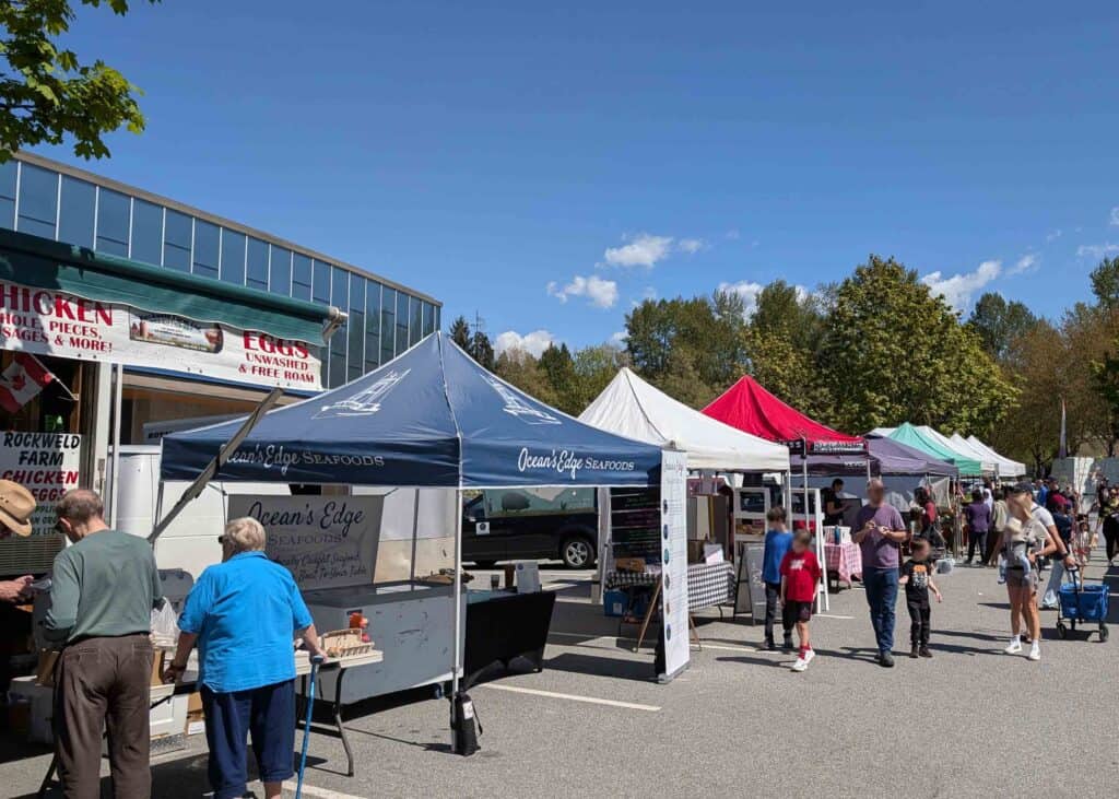 Port Moody Farmers Market with rows of vendor tents selling local goods, people browsing, and stalls offering produce, seafood, and artisan items.