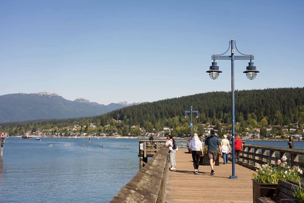 People walking along a wooden pier at Rocky Point Park in Port Moody, with views over Burrard Inlet toward forested hills and distant mountains under a clear blue sky.