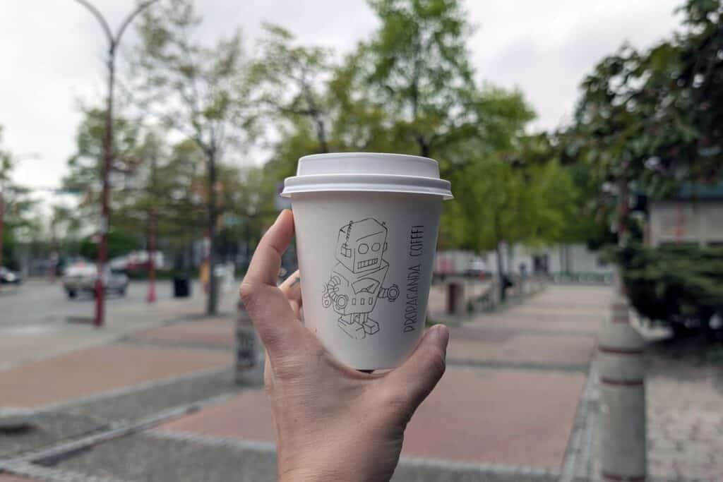 A hand holding a takeaway coffee cup that has a robot drawn on it with the words "Propaganda Coffee."