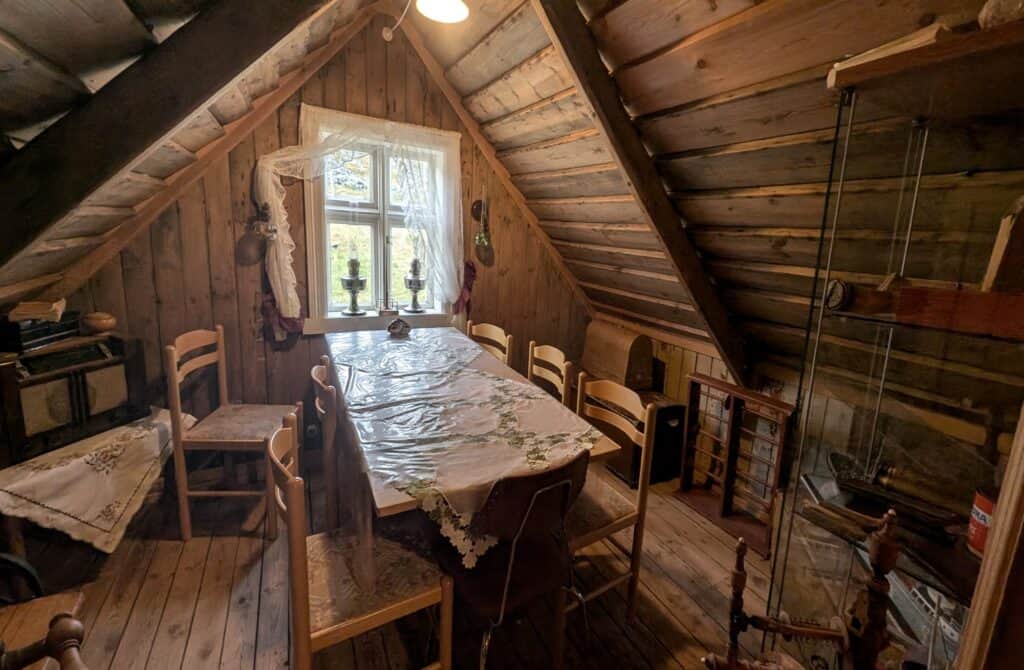 A rustic wooden room inside a historic Icelandic turf house features a dining table, simple wooden chairs, lace curtains, and period furnishings beneath a steeply sloped timber roof.