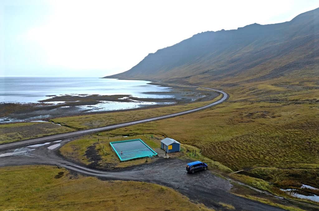 A small geothermal pool sits beside a remote coastal road in Iceland’s Westfjords, with mountains, shoreline, and open water stretching into the distance.