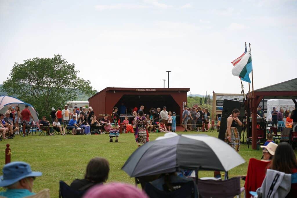 Spectators watch dancers performing in the arena during the Miawpukek First Nation Powwow, with community members gathered around the field.