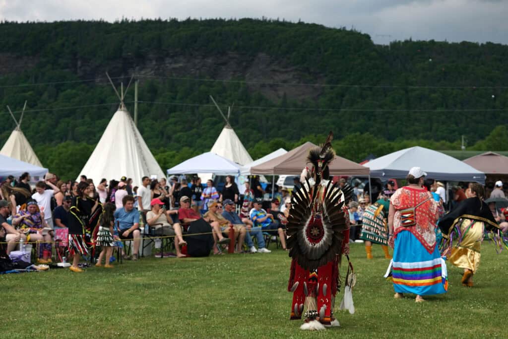 Dancers in colourful regalia taking part in the Grand Entry at the Miawpukek First Nation Powwow, with spectators and traditional tipis in the background.