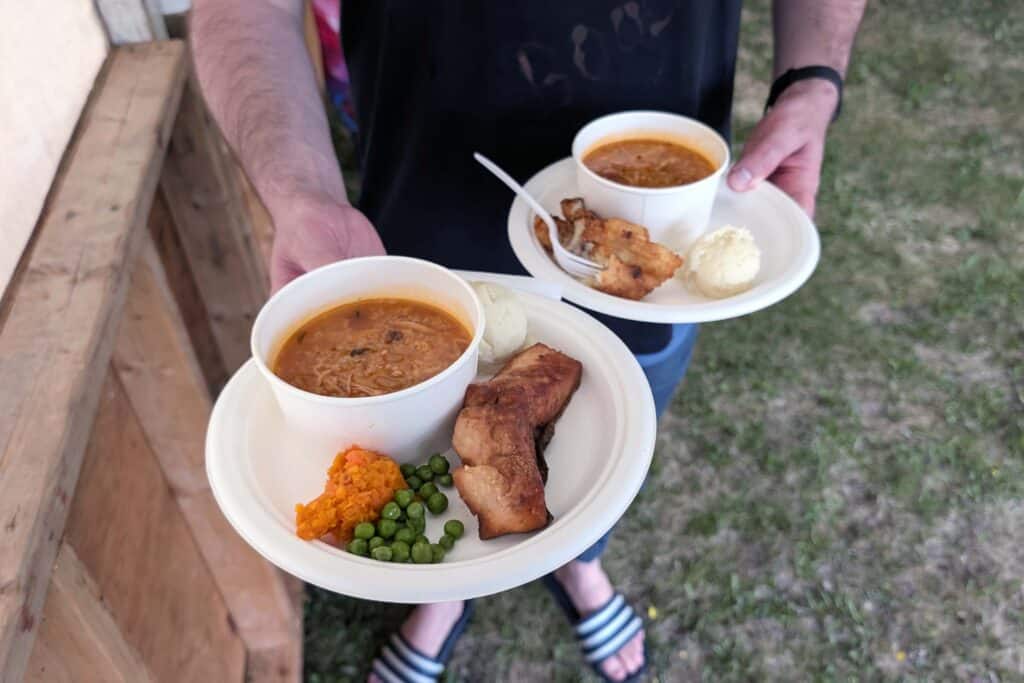 Two white plates with a bowl of soup, salmon, potato, carrot, and peas on them, being held by a man's hands.