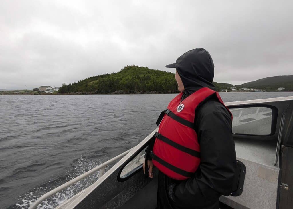 Boat ride across Hare Bay to The Beaches Archaeological Site in Newfoundland, with forested islands and rugged coastline in the background.