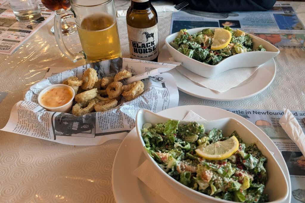 Two bowls of caesar salad and a basket of calamari along with a beer, all presented on a table.