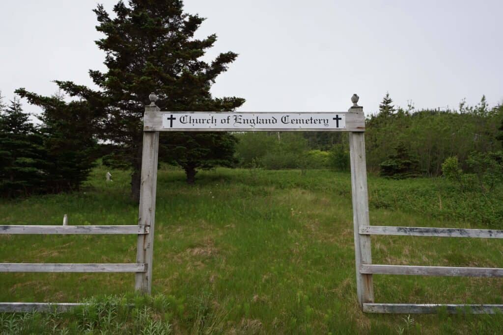 Entrance to the historic Church of England Cemetery in Harbour Breton, Newfoundland, marked by a wooden gateway and surrounded by grassy grounds and evergreen trees.