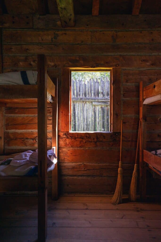 Interior of a reconstructed wooden living quarters at Fort William Historical Park, with bunk beds, brooms and sunlight coming through the window.